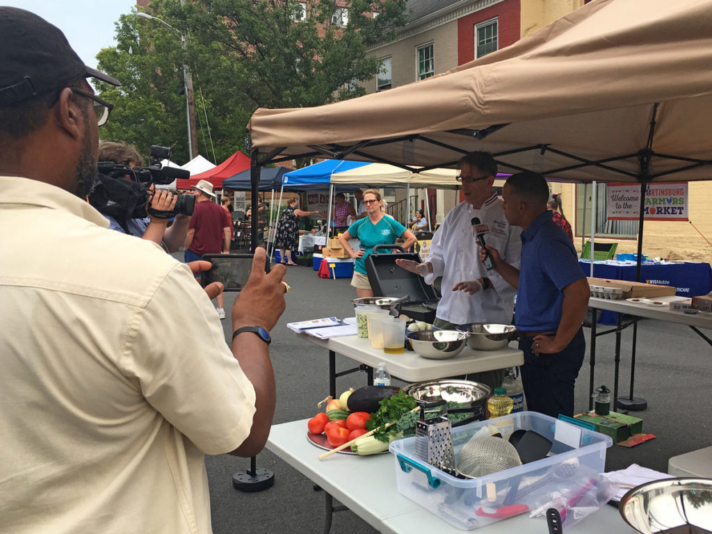 Chef Steve Weiss showing market-goers how to make eggplant bruschetta while being interviewed by the local news stations.